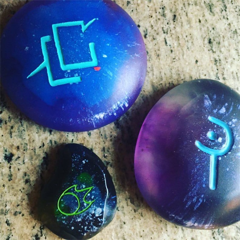 May include: Three round, smooth, painted stones with different symbols. The stones are painted in shades of blue, purple, and black with white speckles. The symbols are painted in turquoise, green, and blue.