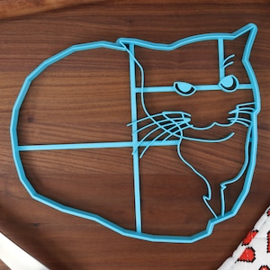 May include: A blue plastic cookie cutter shaped like a cat with a grumpy expression. The cutter is on a wooden surface.