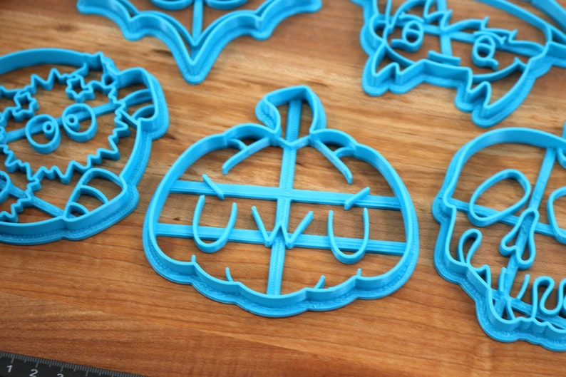 May include: A set of blue plastic cookie cutters shaped like a pumpkin, a skull, a bat, a monster, and a creature with a flower on its head. The pumpkin cutter has the word "uwu" inside.