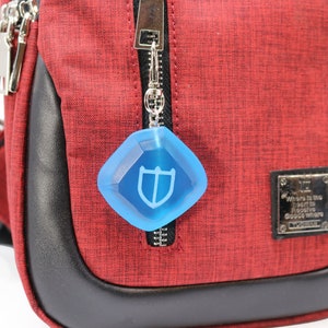 Puede incluir: Un bolso bandolera rojo con una cremallera negra y un llavero azul en forma de diamante con un logotipo de escudo. El bolso tiene un ribete de cuero negro y una etiqueta de metal plateado que dice "D&oacute;nde est&aacute; el coraz&oacute;n para encontrar el lugar donde nacen los sue&ntilde;os."