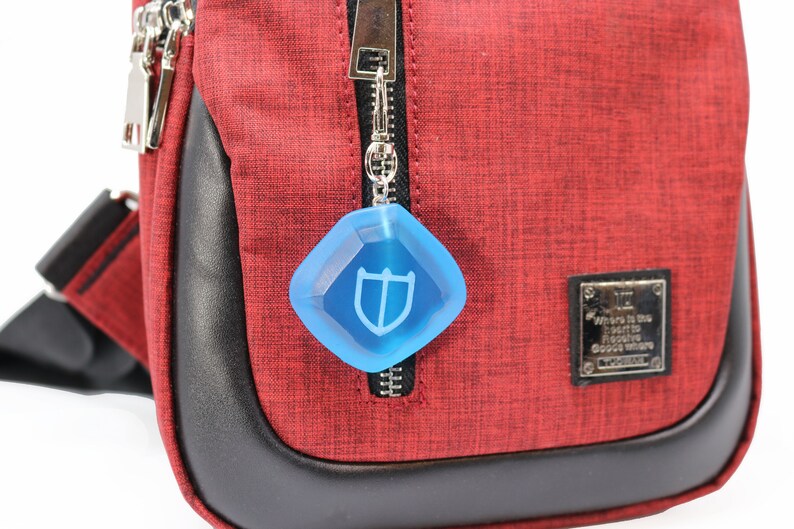 Puede incluir: Un bolso bandolera rojo con una cremallera negra y un llavero azul en forma de diamante con un logotipo de escudo. El bolso tiene un ribete de cuero negro y una etiqueta de metal plateado que dice "D&oacute;nde est&aacute; el coraz&oacute;n para encontrar el lugar donde nacen los sue&ntilde;os."