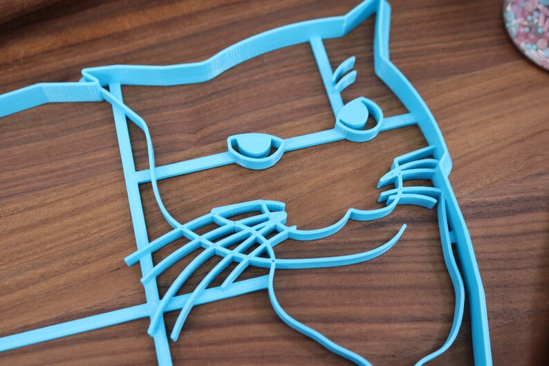 May include: A light blue plastic cookie cutter shaped like a cat's face. The cutter has detailed features, including eyes, a nose, and whiskers.