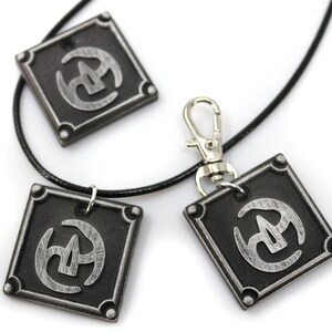 May include: Three silver-coloured pendants with a black and silver symbol on a black cord necklace. The pendants are square shaped with rounded corners and have a silver clasp.