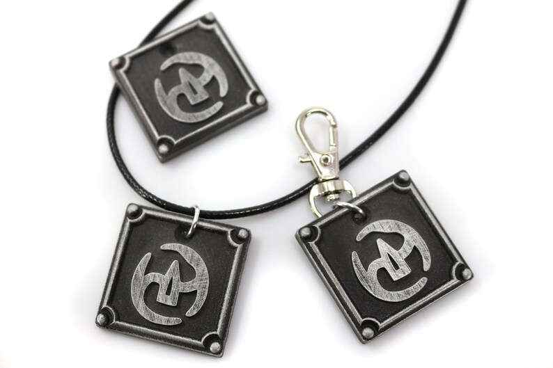 May include: Three silver-coloured pendants with a black and silver symbol on a black cord necklace. The pendants are square shaped with rounded corners and have a silver clasp.