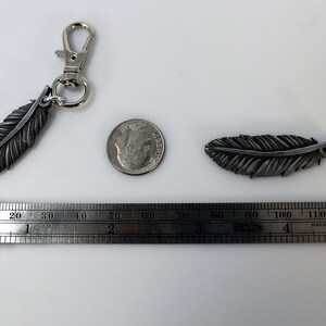 Op de afbeelding: Een zilverkleurige veersleutelhanger met een karabijnhaak, een losse veersier, een dime en een liniaal. De veersleutelhanger is ongeveer 7,6 cm lang. De losse veer is ongeveer 5 cm lang. De liniaal is in inches gemarkeerd.