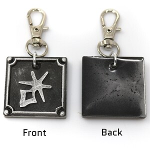 May include: A silver keychain with a black square charm. The front of the charm has a silver kanji character. The back of the charm is plain.
