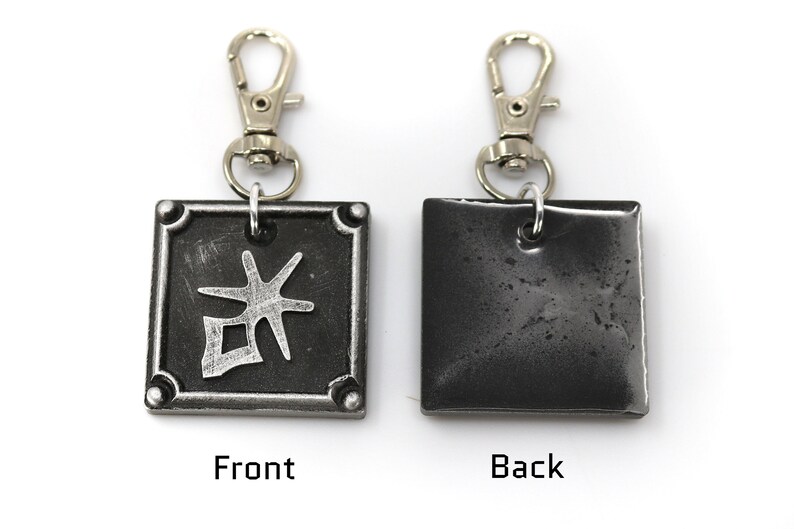 May include: A silver keychain with a black square charm. The front of the charm has a silver kanji character. The back of the charm is plain.