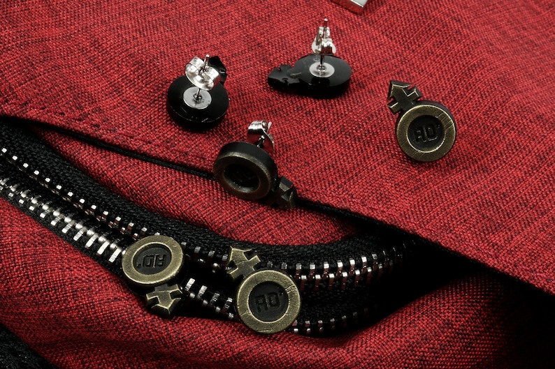 Puede incluir: Un conjunto de seis pendientes con un disco negro con un poste plateado y un s&iacute;mbolo negro y dorado con las letras "RO" sobre un fondo de tela roja.