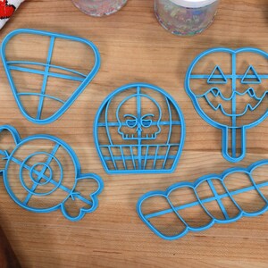 May include: A set of six blue plastic cookie cutters shaped like a candy cane, a skull, a lollipop, a triangle, a swirl, and a candy. The cutters are on a wooden surface.