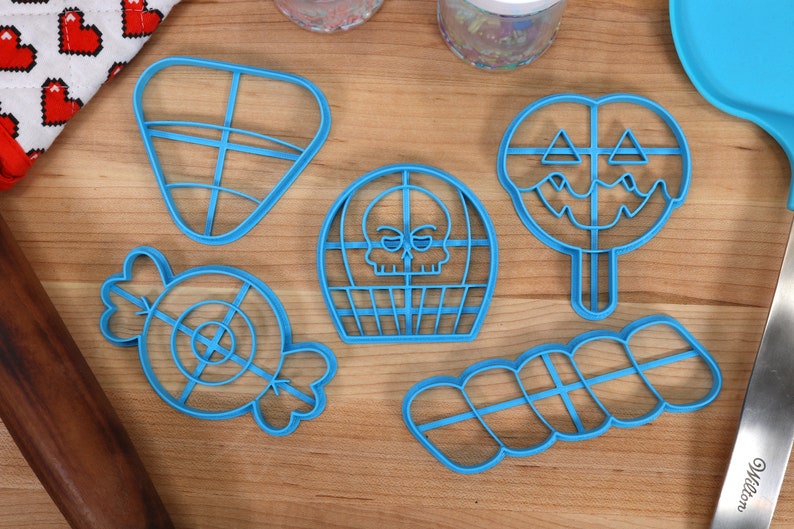 May include: A set of six blue plastic cookie cutters shaped like a candy cane, a skull, a lollipop, a triangle, a swirl, and a candy. The cutters are on a wooden surface.