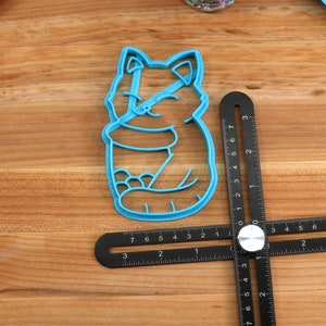 May include: A blue plastic cookie cutter shaped like a cartoon cat sitting with its paws tucked in. The cutter is on a wooden surface with a black ruler.