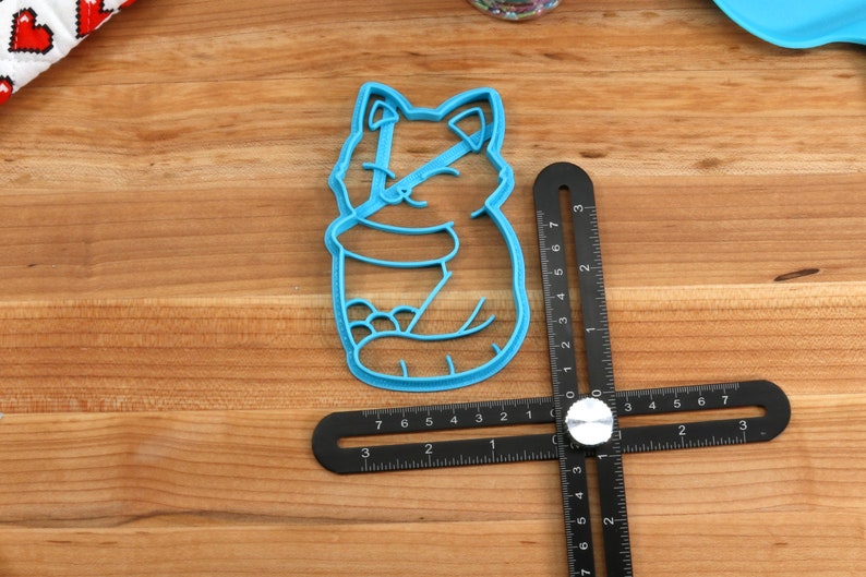 May include: A blue plastic cookie cutter shaped like a cartoon cat sitting with its paws tucked in. The cutter is on a wooden surface with a black ruler.