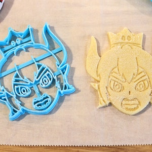 May include: Two cookie cutters, one blue and one yellow, shaped like a cartoon character with a crown, horns, and an angry expression.