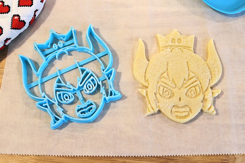 May include: Two cookie cutters, one blue and one yellow, shaped like a cartoon character with a crown, horns, and an angry expression.