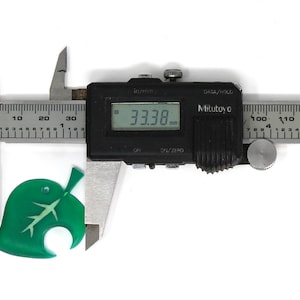 May include: A green leaf-shaped charm with a white stem and veins, measured with a digital caliper showing a measurement of 33.38 mm.