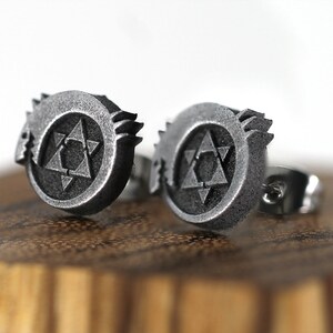 May include: A pair of silver earrings featuring a stylized symbol with a star inside a circle. The symbol is raised and textured.