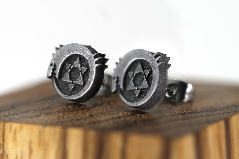 May include: A pair of silver earrings featuring a stylized symbol with a star inside a circle. The symbol is raised and textured.