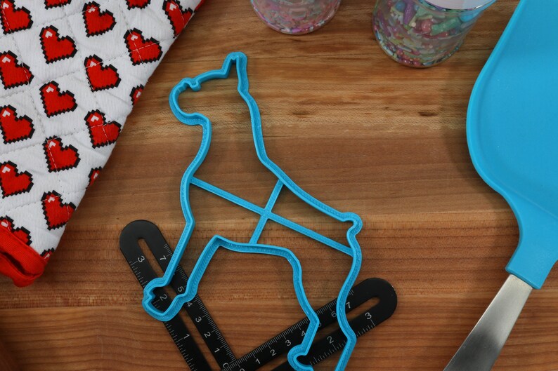 May include: A blue plastic cookie cutter shaped like a dog, on a wooden surface with a ruler and a blue spatula.