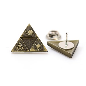 May include: A gold-colored triangular pin with a dark brown design featuring a stylized triforce symbol and stars. The back of the pin has a silver-colored pin back.