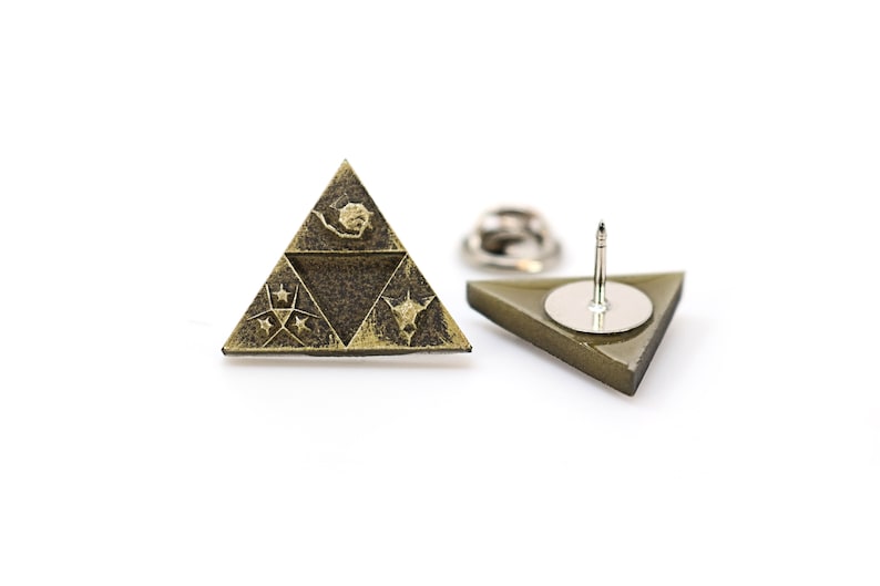May include: A gold-colored triangular pin with a dark brown design featuring a stylized triforce symbol and stars. The back of the pin has a silver-colored pin back.