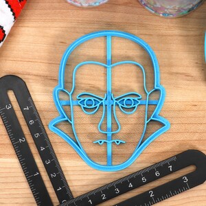 May include: A light blue plastic cookie cutter in the shape of a face with a stern expression. The cutter is on a wooden surface, next to a black ruler with measurements in inches and a white cloth with red pixelated hearts.