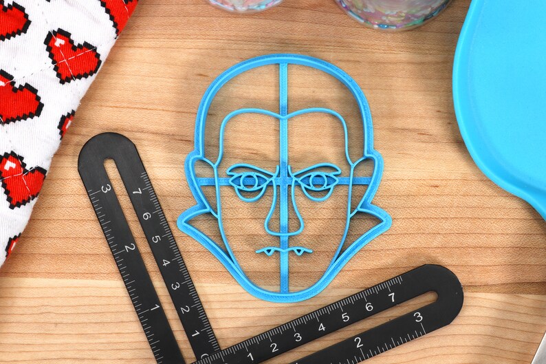 May include: A light blue plastic cookie cutter in the shape of a face with a stern expression. The cutter is on a wooden surface, next to a black ruler with measurements in inches and a white cloth with red pixelated hearts.