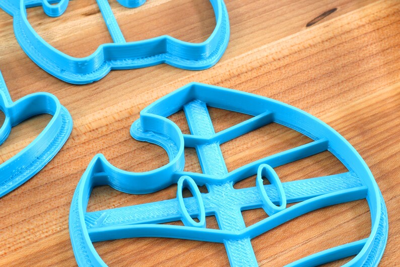 May include: Three blue plastic cookie cutters in the shape of a ghost, a snowman, and a Christmas tree. The cutters are on a wooden surface.