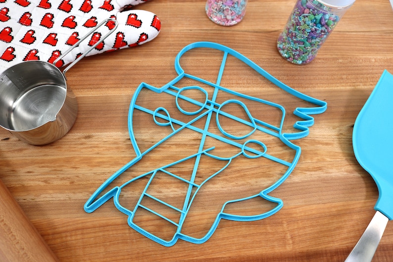 May include: A blue plastic cookie cutter shaped like a cartoon character with a hat. The cutter has a detailed design with lines and curves.