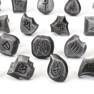 Puede incluir: Una colecci&oacute;n de piedras r&uacute;nicas de metal gris desgastado con s&iacute;mbolos grabados. Las piedras var&iacute;an en forma, incluyendo formas cuadradas, redondas e irregulares. Las runas probablemente se utilizan para la adivinaci&oacute;n o los juegos.