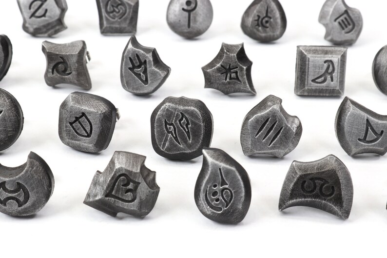 Puede incluir: Una colecci&oacute;n de piedras r&uacute;nicas de metal gris desgastado con s&iacute;mbolos grabados. Las piedras var&iacute;an en forma, incluyendo formas cuadradas, redondas e irregulares. Las runas probablemente se utilizan para la adivinaci&oacute;n o los juegos.