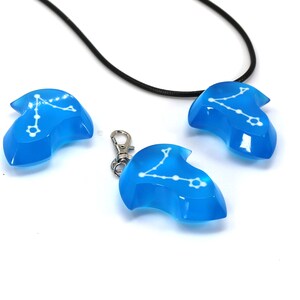 K&ouml;nnte beinhalten: Drei blaue Harz-Anh&auml;nger mit einem wei&szlig;en Sternbild-Design. Ein Anh&auml;nger ist an einem schwarzen Band, und ein anderer an einer silbernen Kette.