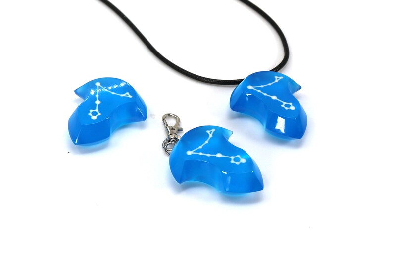K&ouml;nnte beinhalten: Drei blaue Harz-Anh&auml;nger mit einem wei&szlig;en Sternbild-Design. Ein Anh&auml;nger ist an einem schwarzen Band, und ein anderer an einer silbernen Kette.