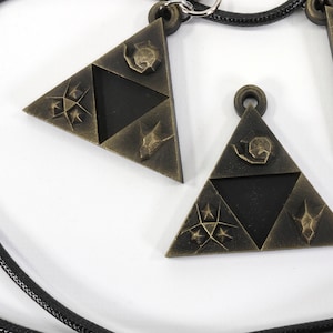 May include: A black cord necklace with three gold-toned triangular pendants. Each pendant features a stylized design of a triangle with a symbol inside.