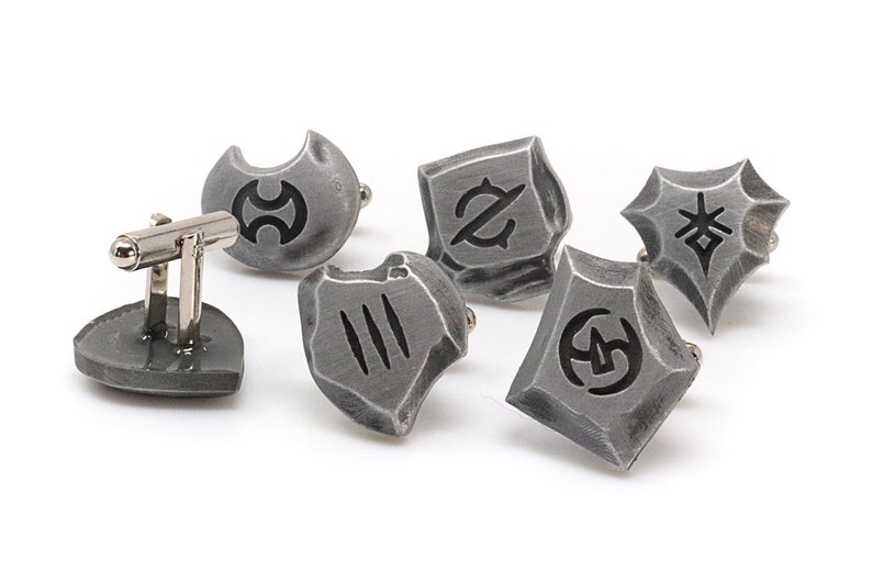 May include: Set of six silver cufflinks with different black symbols on each. The cufflinks are shaped like shields and have a textured surface.