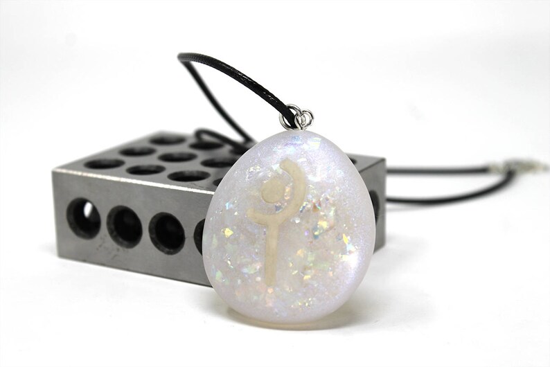 K&ouml;nnte beinhalten: Eine wei&szlig;e, irisierende Halskette mit einem silbernen Kettenband und einem wei&szlig;en Symbol auf dem Anh&auml;nger. Der Anh&auml;nger ist oval geformt und hat eine glitzernde Oberfl&auml;che.