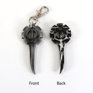 May include: A silver-colored metal keychain with a circular design on one side and a pointed tip on the other. The circular design features a detailed pattern and a small hole in the center. The keychain has a silver-colored metal loop for attaching to a key ring.