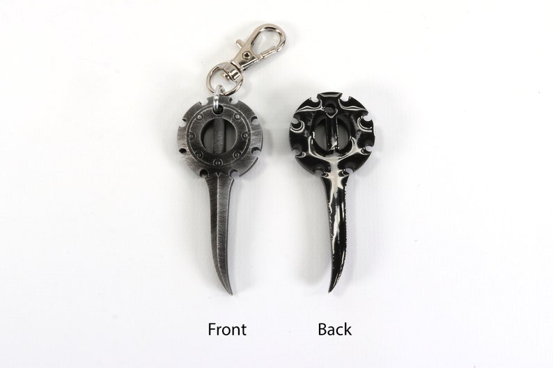 May include: A silver-colored metal keychain with a circular design on one side and a pointed tip on the other. The circular design features a detailed pattern and a small hole in the center. The keychain has a silver-colored metal loop for attaching to a key ring.