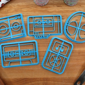 May include: Five blue plastic cookie cutters shaped like retro music devices: a record player, a cassette tape, a pair of headphones, a cassette tape, and a boombox.