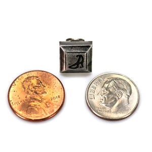 May include: A silver-coloured metal charm with a black letter "A" in the centre. The charm is square-shaped and has a raised border. The charm is shown next to two UK coins, a penny and a dime, for size comparison.