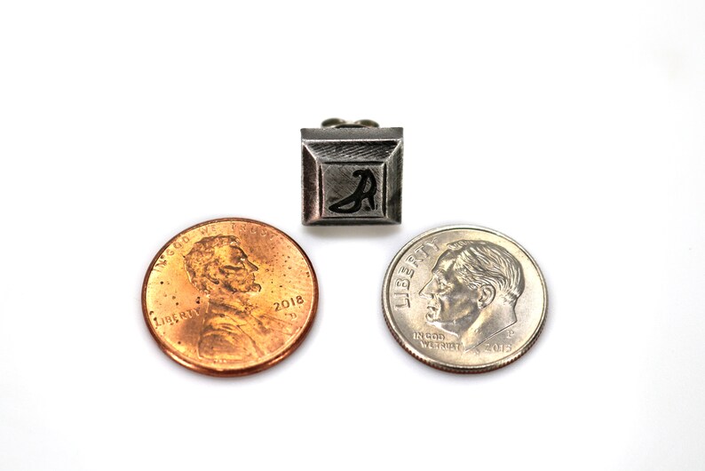 May include: A silver-coloured metal charm with a black letter "A" in the centre. The charm is square-shaped and has a raised border. The charm is shown next to two UK coins, a penny and a dime, for size comparison.
