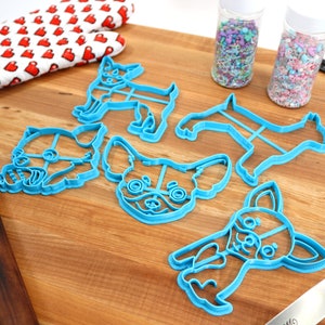 Puede incluir: Un juego de cinco cortadores de galletas de pl&aacute;stico azul con forma de diferentes variaciones de un perro chihuahua. Los cortadores est&aacute;n sobre una superficie de madera con chispas en frascos y un rodillo.