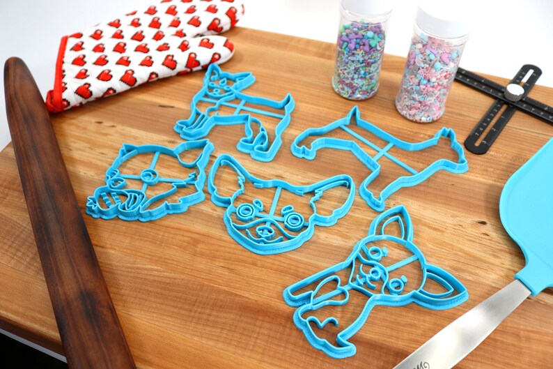 Puede incluir: Un juego de cinco cortadores de galletas de pl&aacute;stico azul con forma de diferentes variaciones de un perro chihuahua. Los cortadores est&aacute;n sobre una superficie de madera con chispas en frascos y un rodillo.