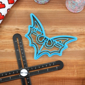 May include: A blue plastic bat cookie cutter on a wooden surface. The cutter has a detailed design with intricate cutouts.