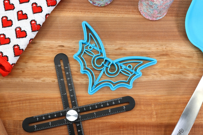 May include: A blue plastic bat cookie cutter on a wooden surface. The cutter has a detailed design with intricate cutouts.