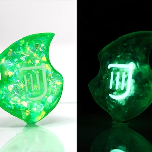 Op de afbeelding: Een groene, iriserende, glow-in-the-dark harsbedel in de vorm van een blad met een wit liersymbool in het midden.