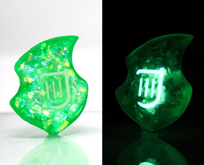Op de afbeelding: Een groene, iriserende, glow-in-the-dark harsbedel in de vorm van een blad met een wit liersymbool in het midden.