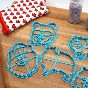 May include: Five blue plastic cookie cutters shaped like Pomeranian dogs. The cutters are on a wooden surface with a rolling pin, a red and white heart patterned oven mitt, and two jars of sprinkles.