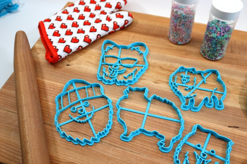 May include: Five blue plastic cookie cutters shaped like Pomeranian dogs. The cutters are on a wooden surface with a rolling pin, a red and white heart patterned oven mitt, and two jars of sprinkles.