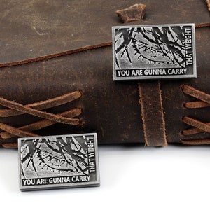 May include: Two silver metal pins with a black and white image of a forest and the text "You are gunna carry that weight".