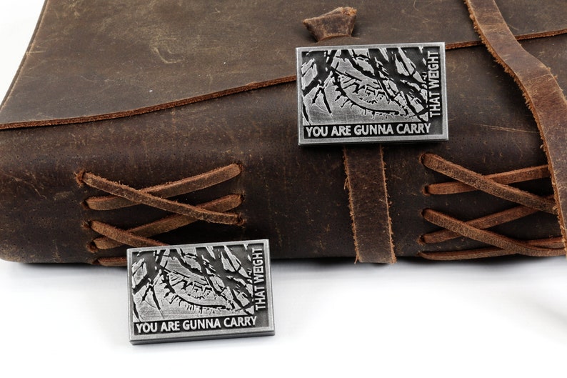 May include: Two silver metal pins with a black and white image of a forest and the text "You are gunna carry that weight".